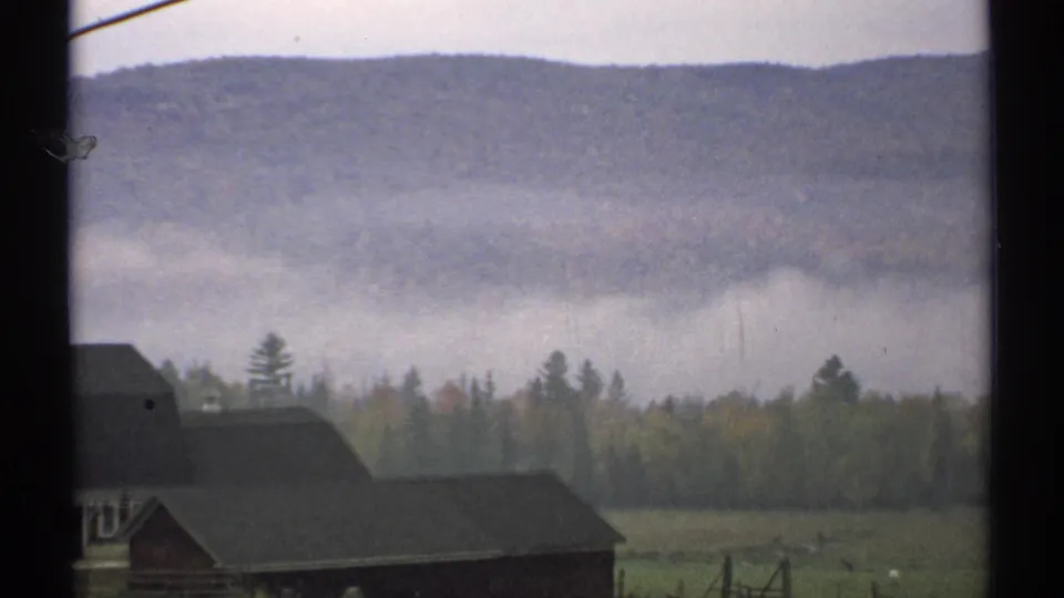 A closer 1971 New Hampshire 8mm view shows dark farm roofs, autumn treeline, low fog, and a wide wooded mountain slope.