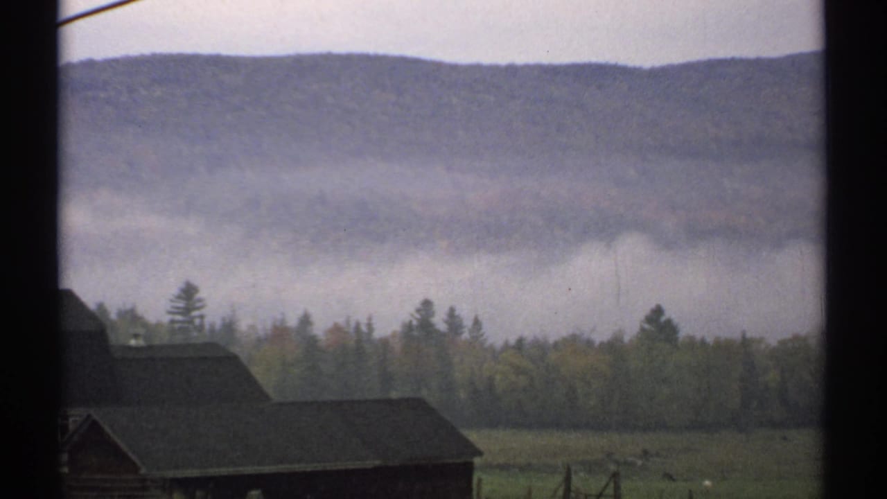 Misty mountain ridge and forest seen through a dark window frame with a barn roof at lower left.