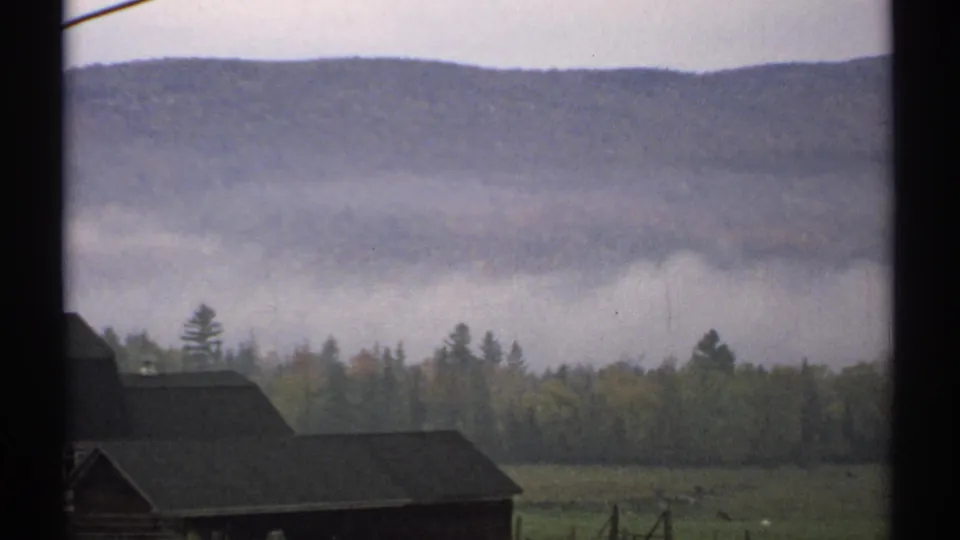 1971 New Hampshire rural landscape with mountain fog, mixed forest, fields, and a barn roof viewed through a window frame.