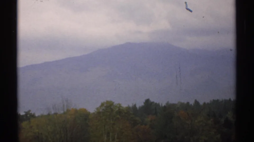 1971 New Hampshire 8mm view of a hazy mountain ridge rising behind a band of forest and low clouds.