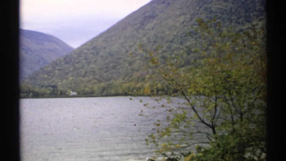 1971 New Hampshire home-movie frame of a lake and wooded mountain slopes, with foliage at the right edge.