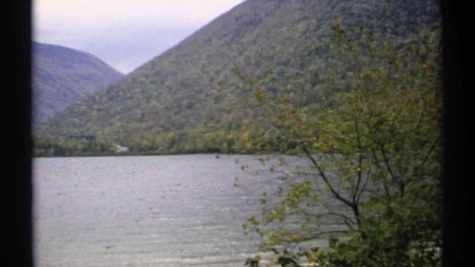 1971 New Hampshire home-movie frame with calm water, forested hills, and shoreline foliage in soft 8mm color.