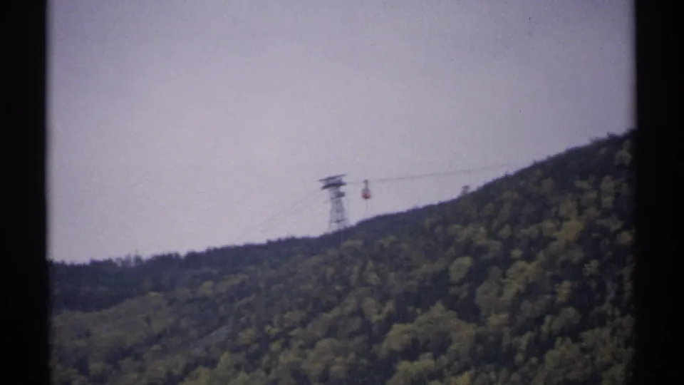 Distant New Hampshire tramway car and steel tower appear above a dense green mountainside in soft 8mm color.