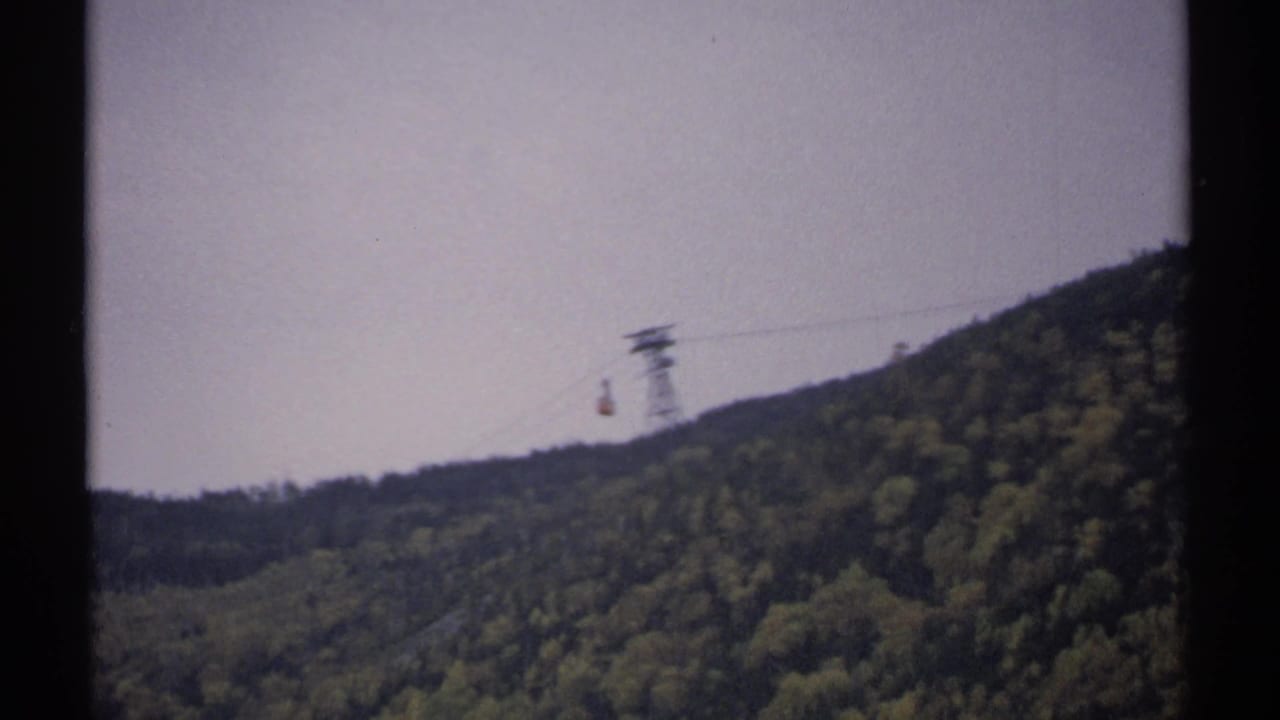 Small tram car hangs from a cable over a green wooded ridge.