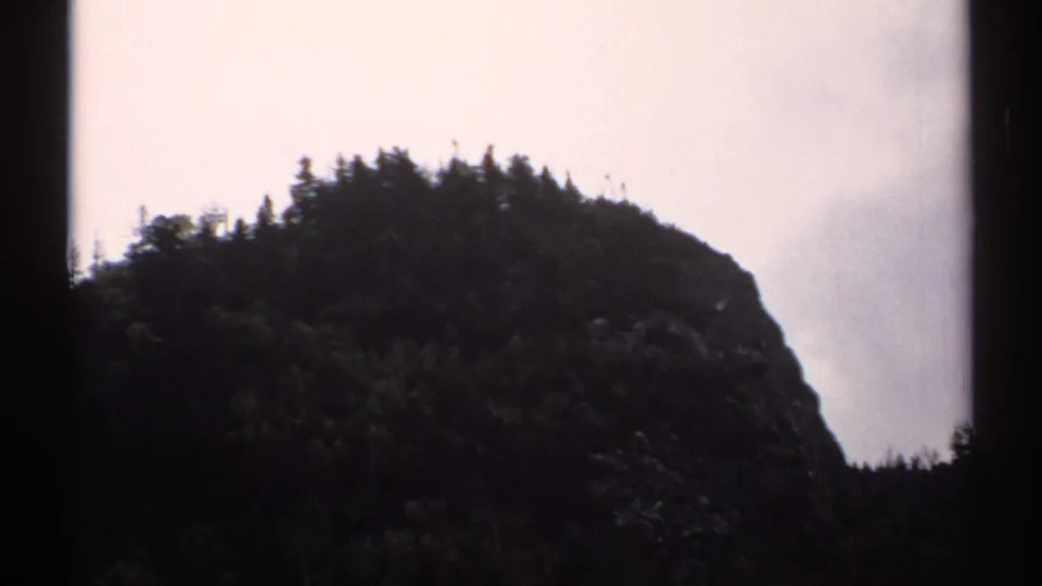 A forested rock face and pale sky appear in a soft 8mm frame from the New Hampshire mountain sequence.