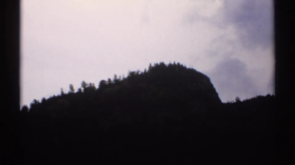 1971 New Hampshire home-movie frame with a dark wooded ridge crossing the lower view under an overcast sky.