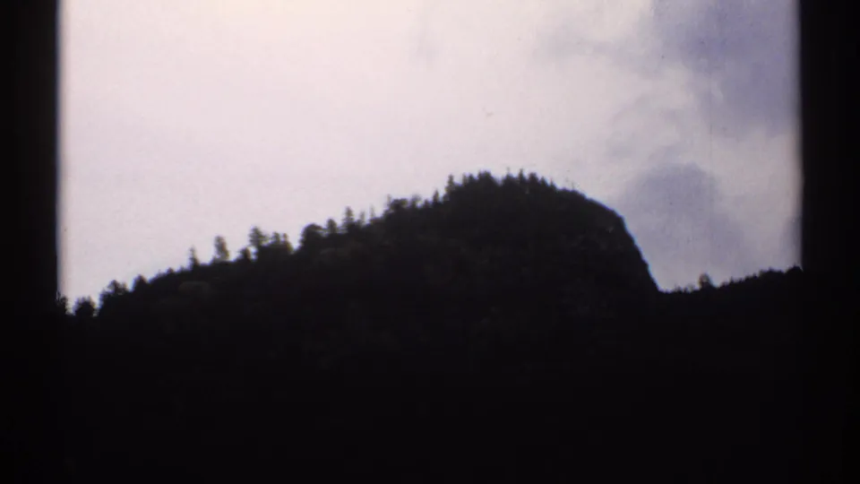 1971 New Hampshire 8mm frame showing a forested mountain ridge in dark silhouette beneath a washed cloudy sky.