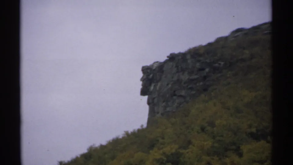 1971 New Hampshire 8mm frame showing a rugged rock outcrop rising over dense green and yellow trees beneath a muted sky.