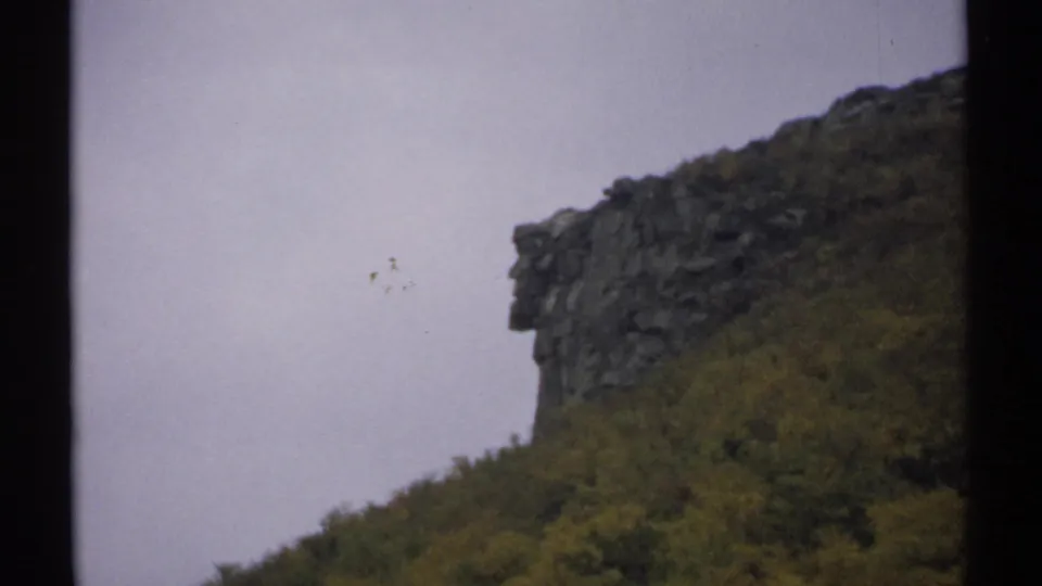 1971 New Hampshire 8mm frame with a rocky mountain edge at upper right, dense hillside vegetation, and a washed pale sky.