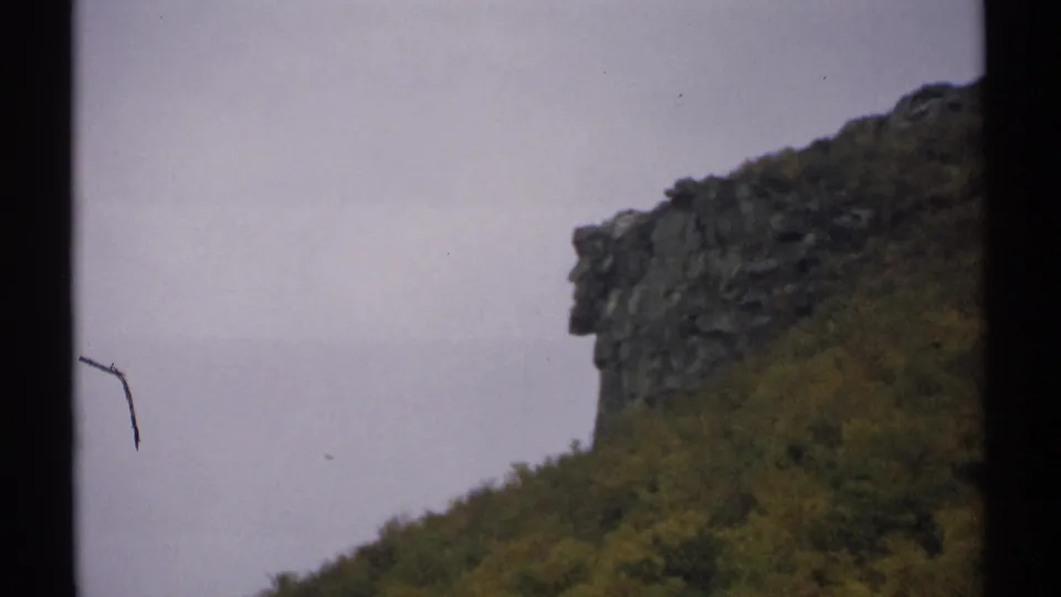 1971 New Hampshire 8mm frame showing a wooded mountain slope and rocky ledge, with visible film border and small surface marks from the scan.