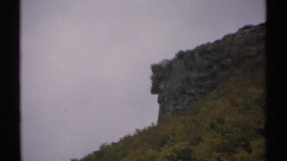 1971 New Hampshire 8mm frame holding on a dark rocky formation above a green and yellow wooded hillside under an overcast sky.