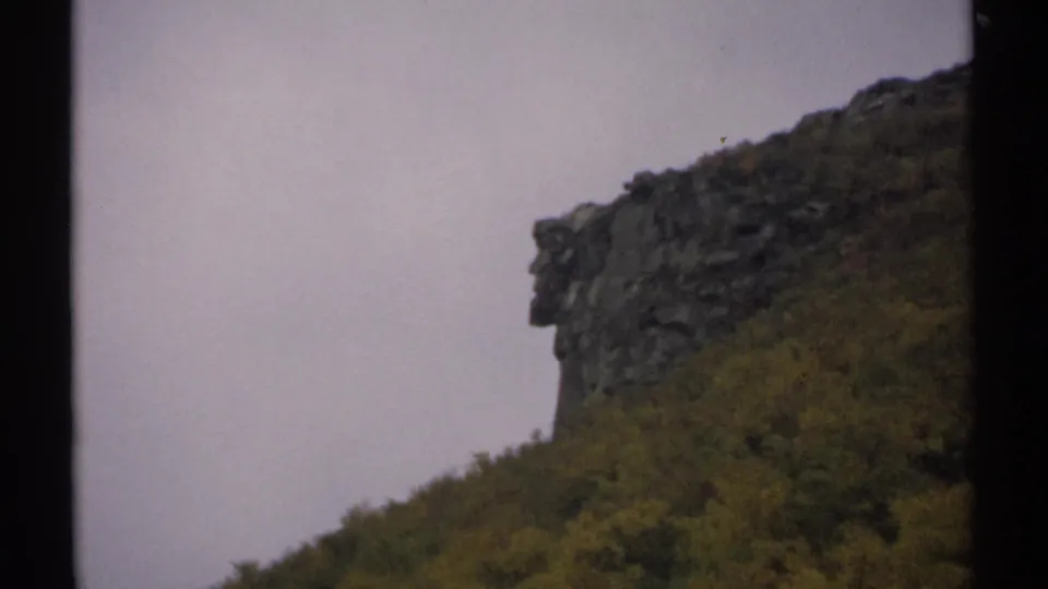 1971 New Hampshire 8mm frame showing a wooded hillside and prominent dark rock outcrop in autumn-toned light.
