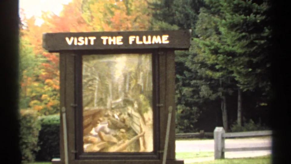 1971 New Hampshire home-movie frame showing the VISIT THE FLUME sign, scenic illustration, colorful foliage, evergreens, and roadside barrier.