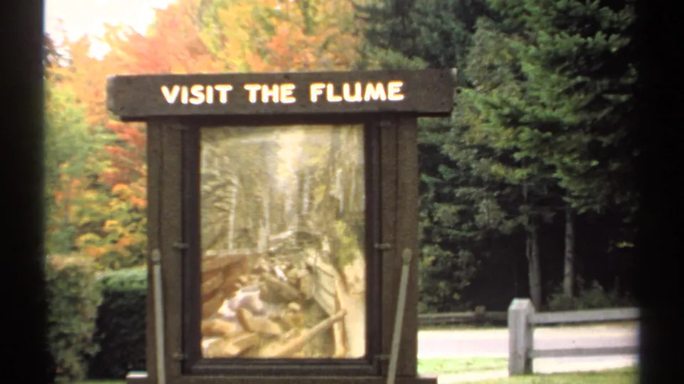 1971 New Hampshire 8mm frame centering the VISIT THE FLUME sign before autumn foliage, evergreens, a pale road edge, and short posts.