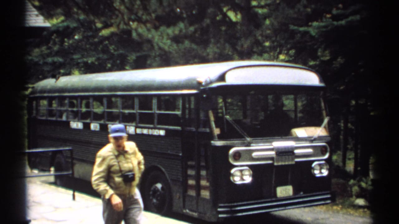 Older man walks in front of a dark bus parked beside trees and a railing.