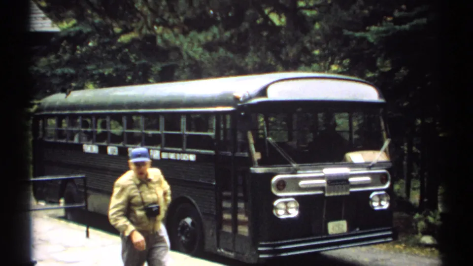 1971 New Hampshire frame with an older man wearing a blue cap and camera walking near a parked tour bus in a wooded area.