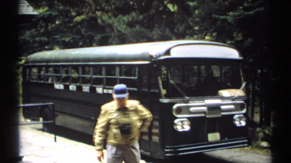 1971 New Hampshire home-movie frame showing a man with a neck camera moving toward the camera with the tour bus behind him.