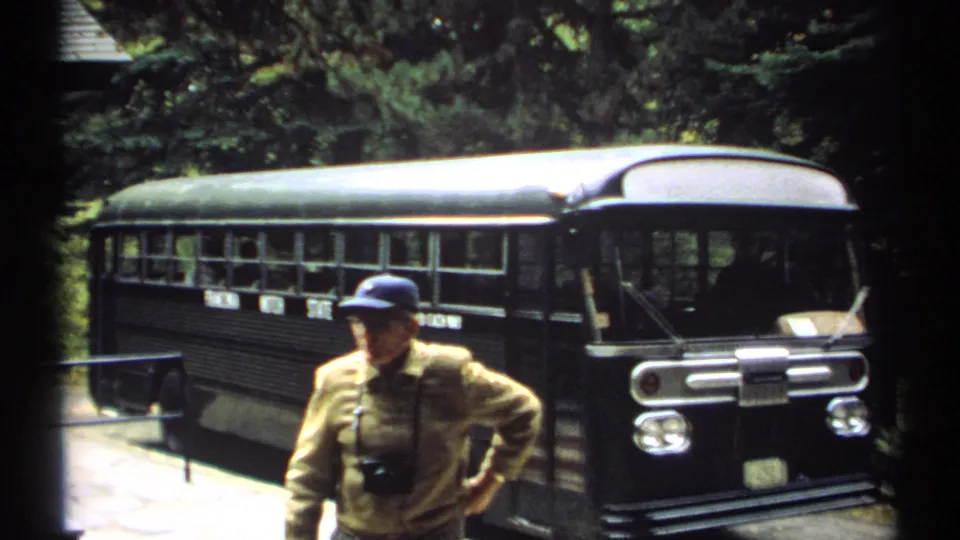 1971 New Hampshire frame showing an older visitor in a blue cap standing before a dark tour bus in a wooded sightseeing stop.