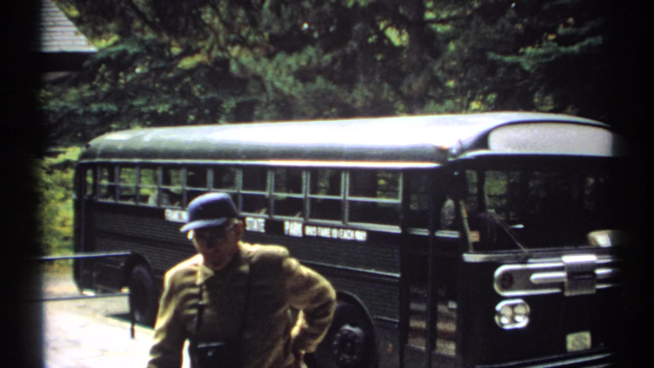 1971 New Hampshire 8mm frame showing an adult man in a blue cap near a black tour bus, with trees and rocky terrain behind him.