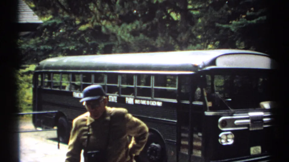 1971 New Hampshire 8mm frame showing an adult man in a blue cap near a black tour bus, with trees and rocky terrain behind him.