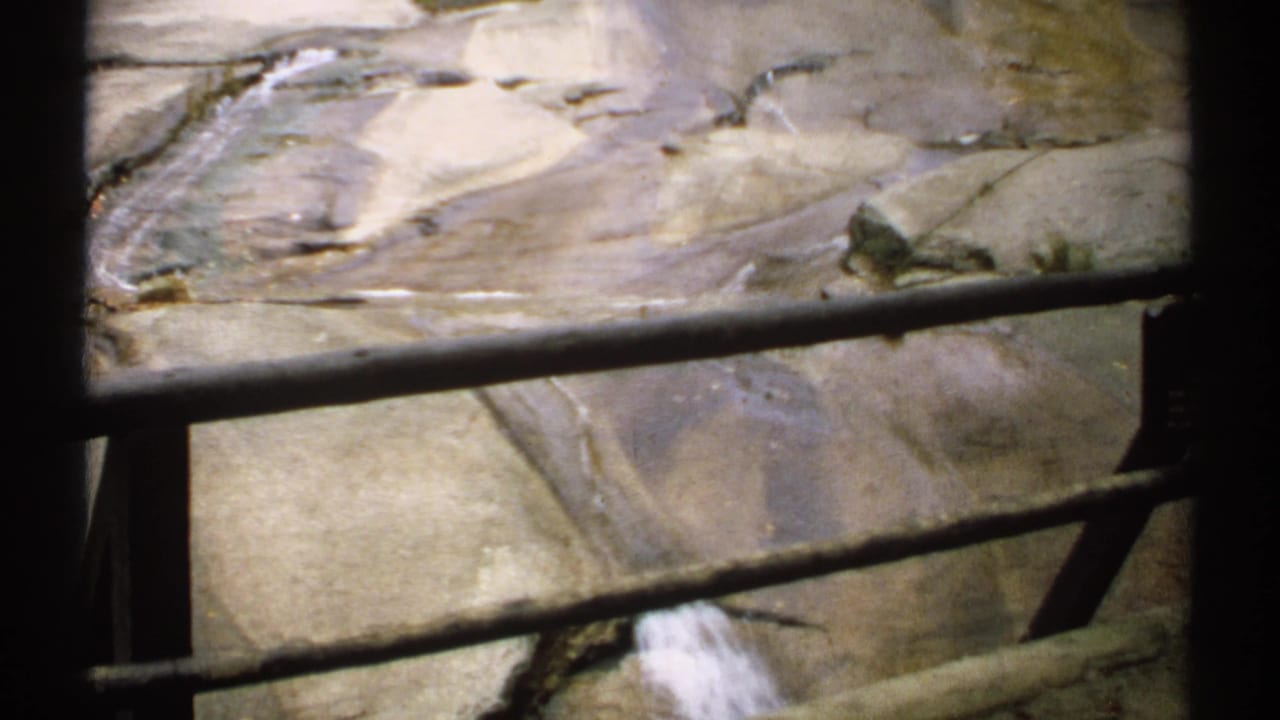 Smooth rock surface and shallow flowing water seen through dark metal railings.