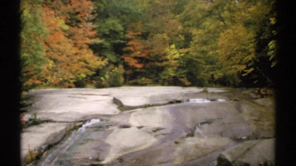 Wide 1971 New Hampshire 8mm view of a rocky stream bordered by autumn trees in orange, yellow, and green tones.