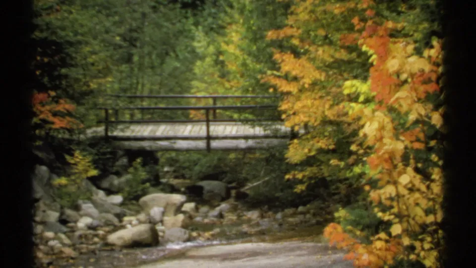 1971 New Hampshire 8mm frame showing a simple wooden bridge over a rocky creek, surrounded by green and yellow fall leaves.