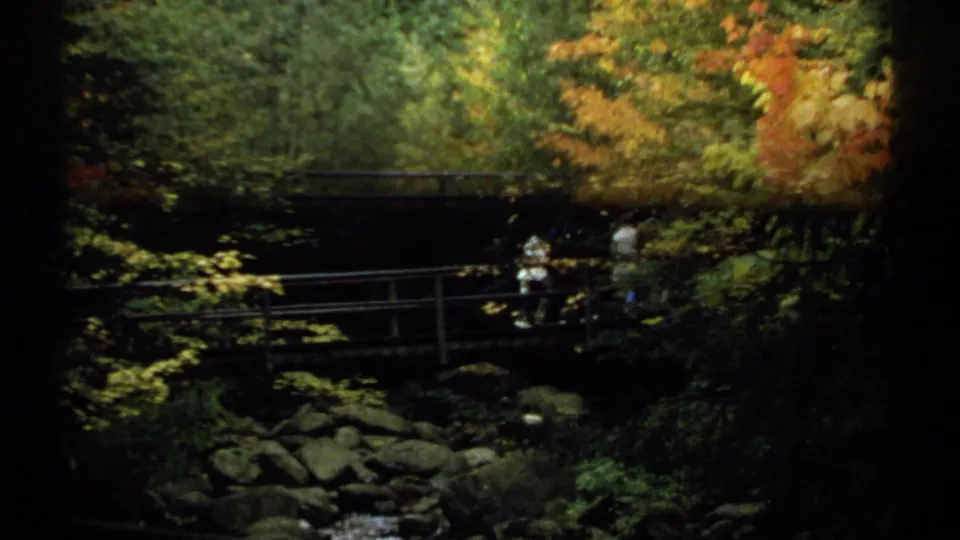 1971 New Hampshire 8mm frame with a distant person in a white shirt crossing a wooden bridge over rocks in a shaded forest.