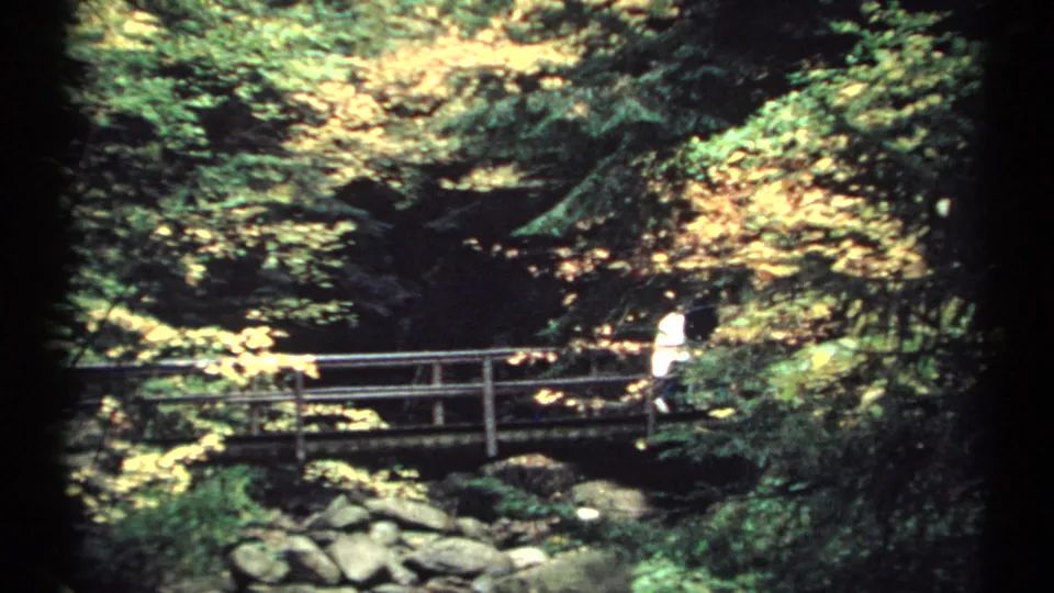 1971 New Hampshire 8mm frame showing a person on a wooden footbridge, partly surrounded by bright yellow foliage and rocks below.