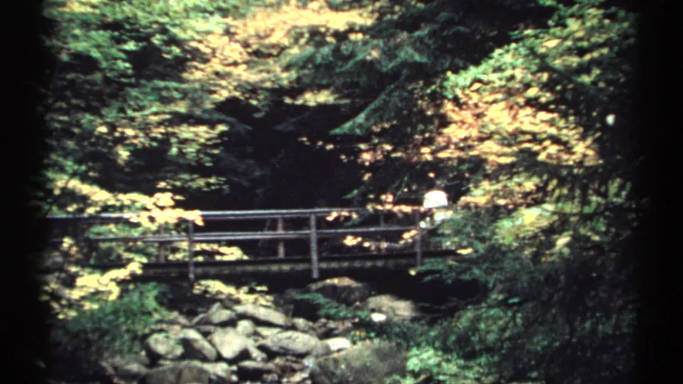 1971 New Hampshire 8mm frame with a distant person near the far side of a wooden bridge over a rocky stream in autumn woods.
