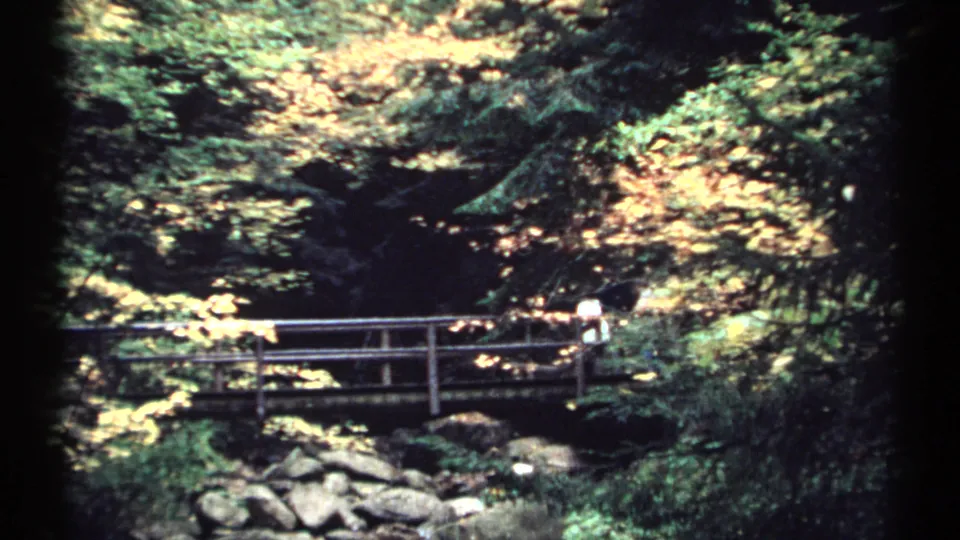 1971 New Hampshire 8mm frame showing a wooden bridge over rocks and water beneath dense trees.