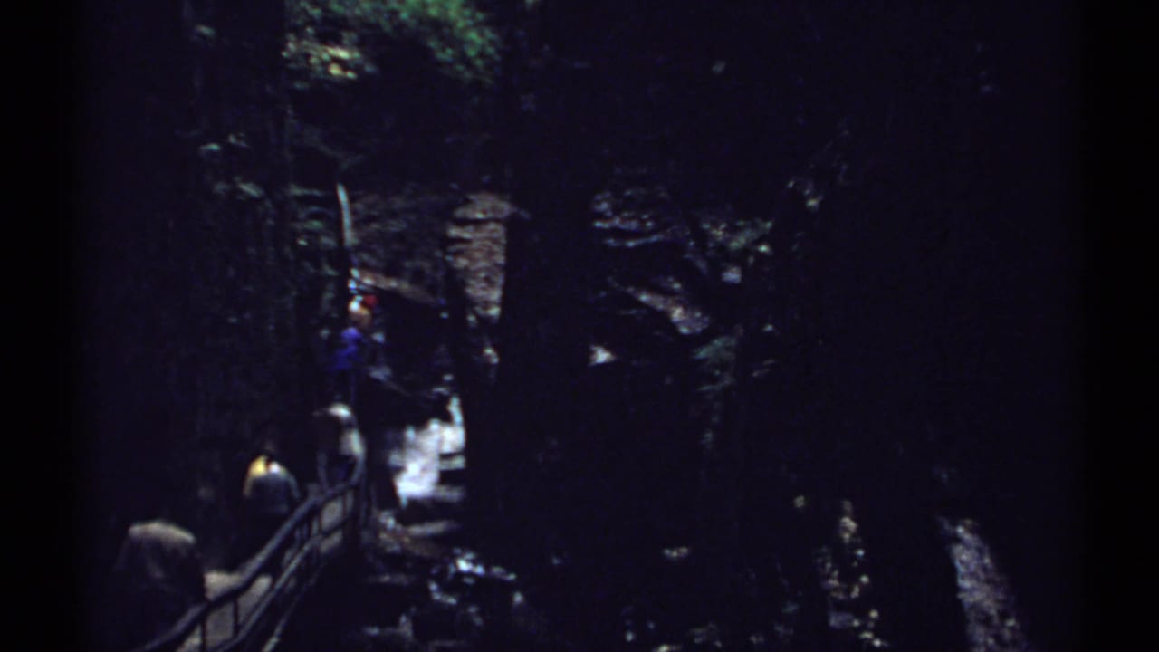 Hikers move along a railing in a dark rocky gorge near falling water.