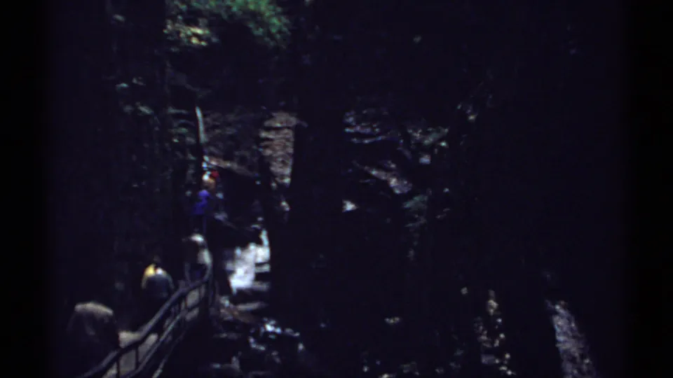 1971 New Hampshire 8mm frame with distant hikers on a railed path inside a shadowed rocky gorge beside water.