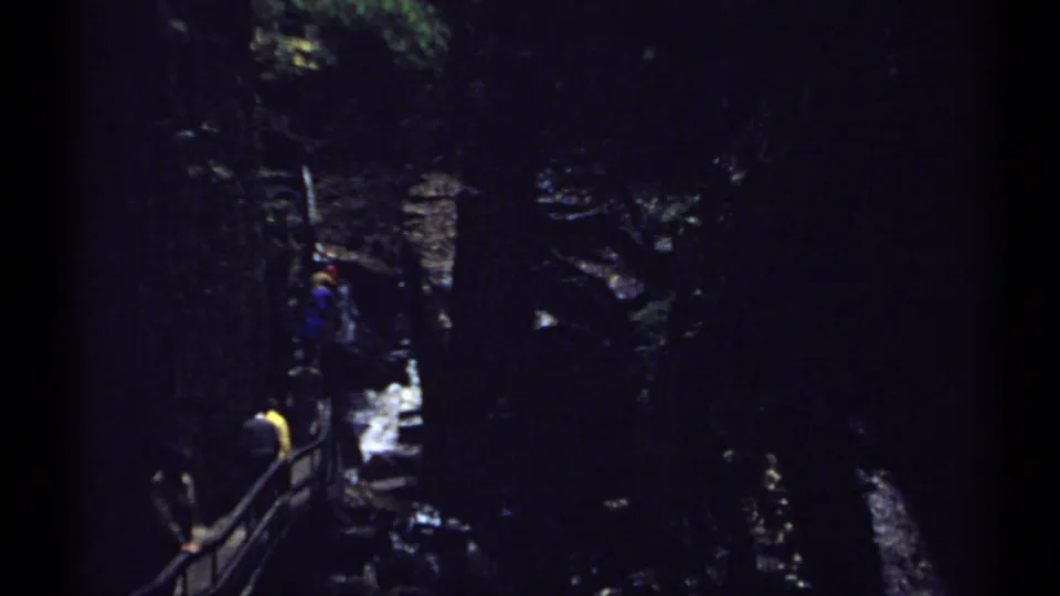 1971 New Hampshire 8mm frame of distant hikers beside a railing in a shadowed canyon near cascading water.