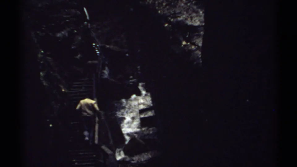 A man climbs farther up the shadowed metal stairs, framed by dark rock, trees, and white water in a New Hampshire ravine.