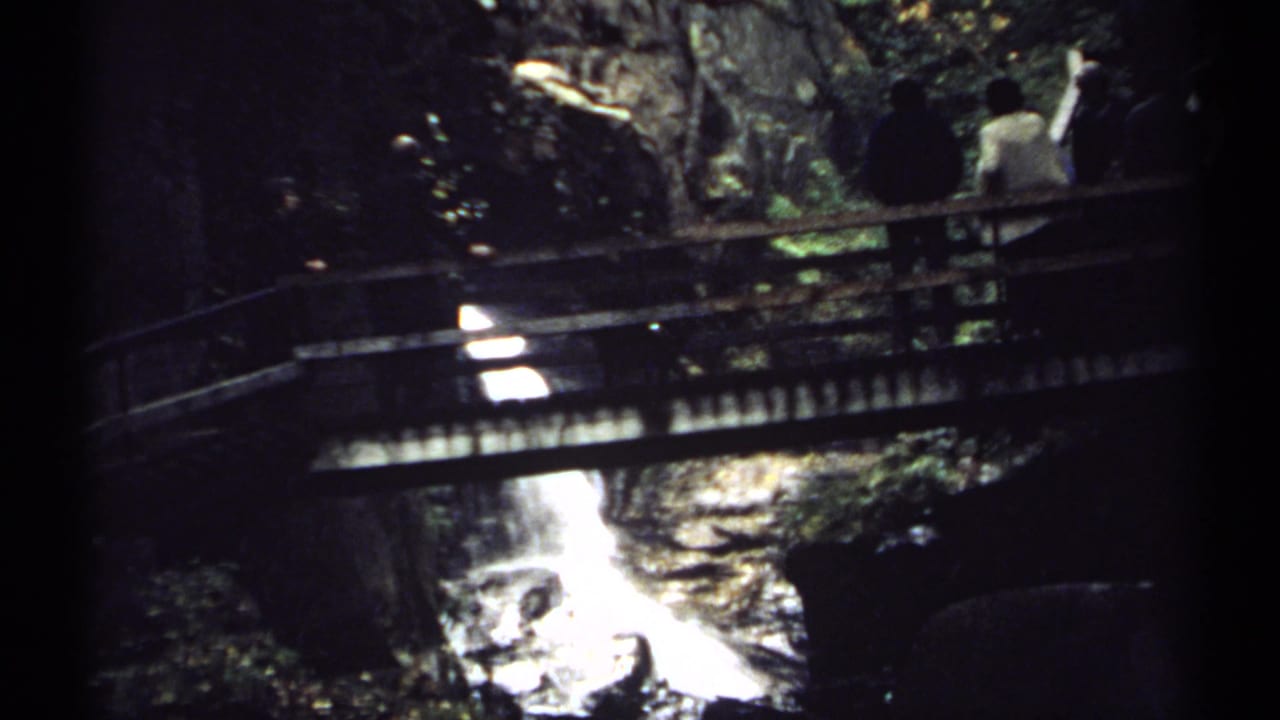 1971 New Hampshire 8mm frame showing visitors on a rustic wooden footbridge over a bright waterfall or fast stream in dense shade.
