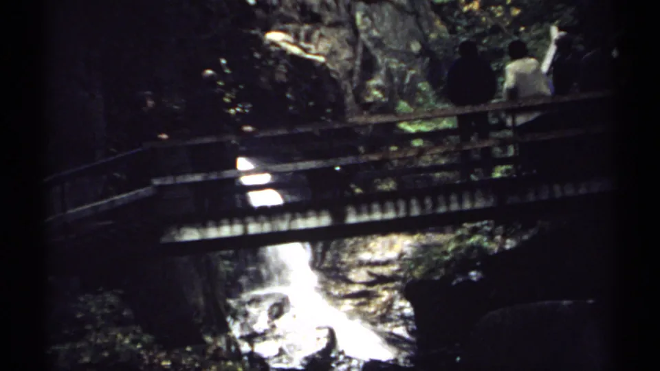 1971 New Hampshire 8mm frame showing visitors on a rustic wooden footbridge over a bright waterfall or fast stream in dense shade.