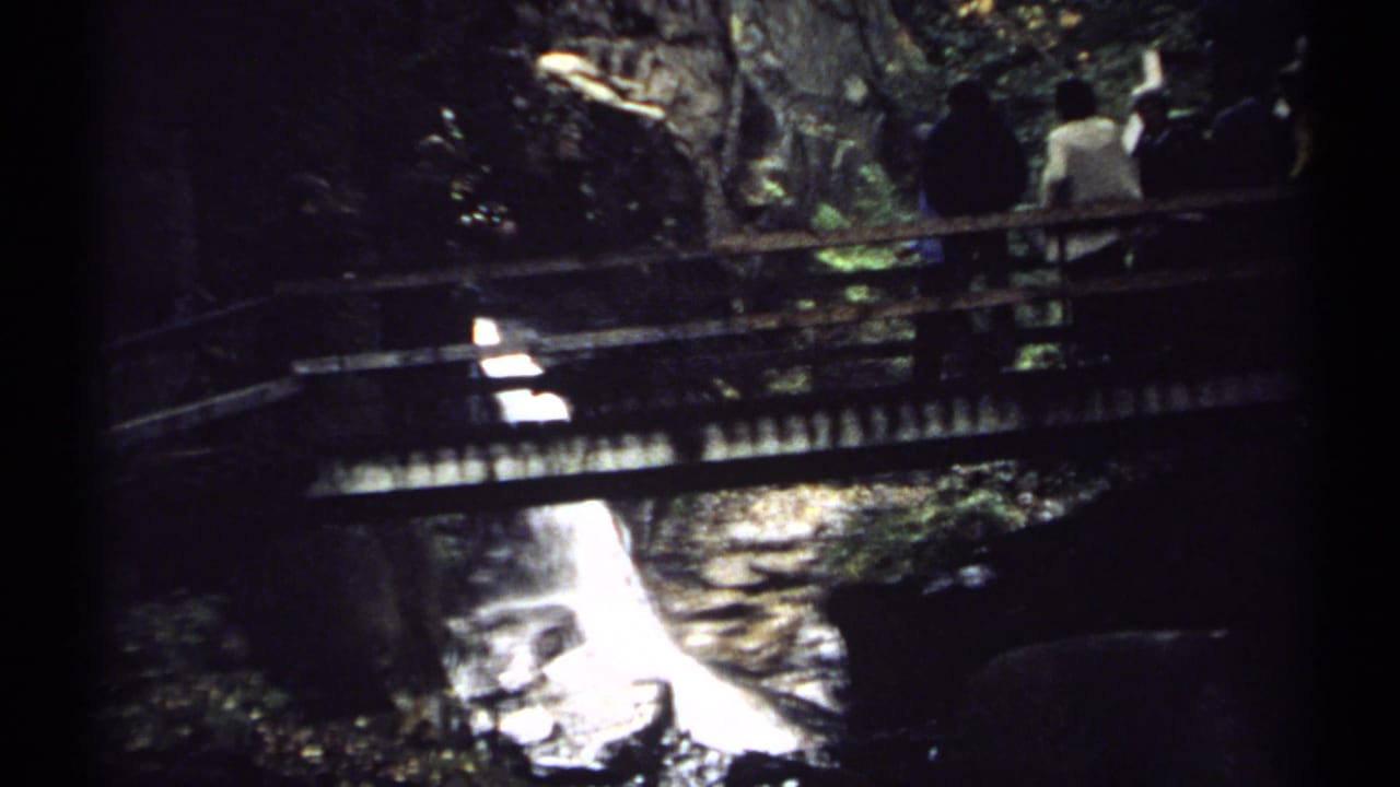 Wooden bridge spans a shaded rocky stream with people visible near the far side.