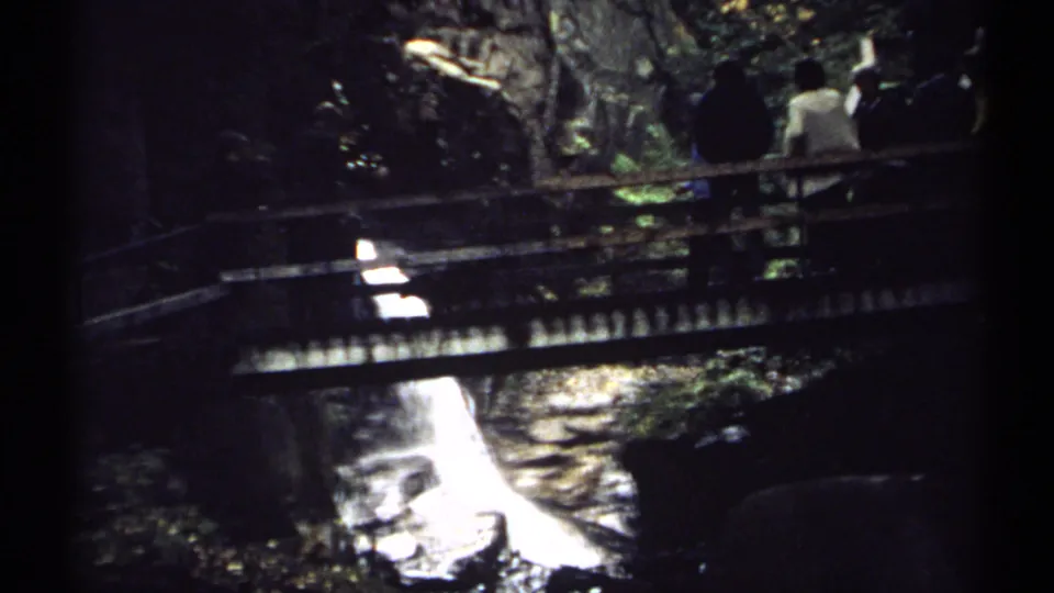 A dark 8mm frame from New Hampshire shows people near a wooden bridge while white water drops through rocks below.