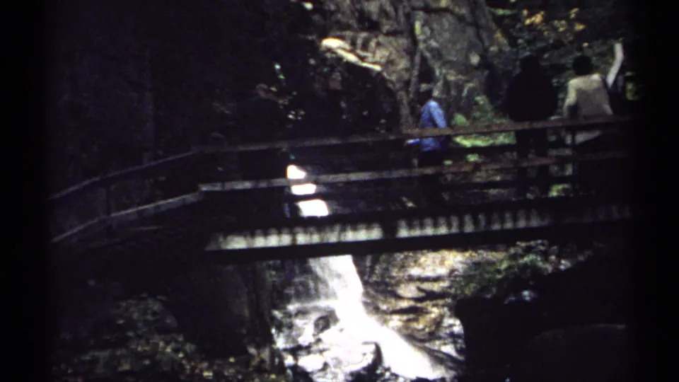 Visitors in a wooded New Hampshire scene cross a rustic bridge above a white cascade, captured with grainy 8mm home-movie texture.