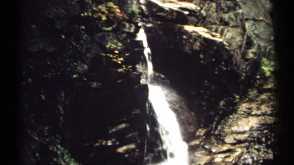 1971 New Hampshire 8mm frame showing a bright narrow waterfall descending over dark rocks beside patches of green foliage.