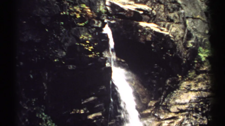 1971 New Hampshire 8mm frame of a bright waterfall running down wet rock, bordered by green leaves and sunlit stone surfaces.