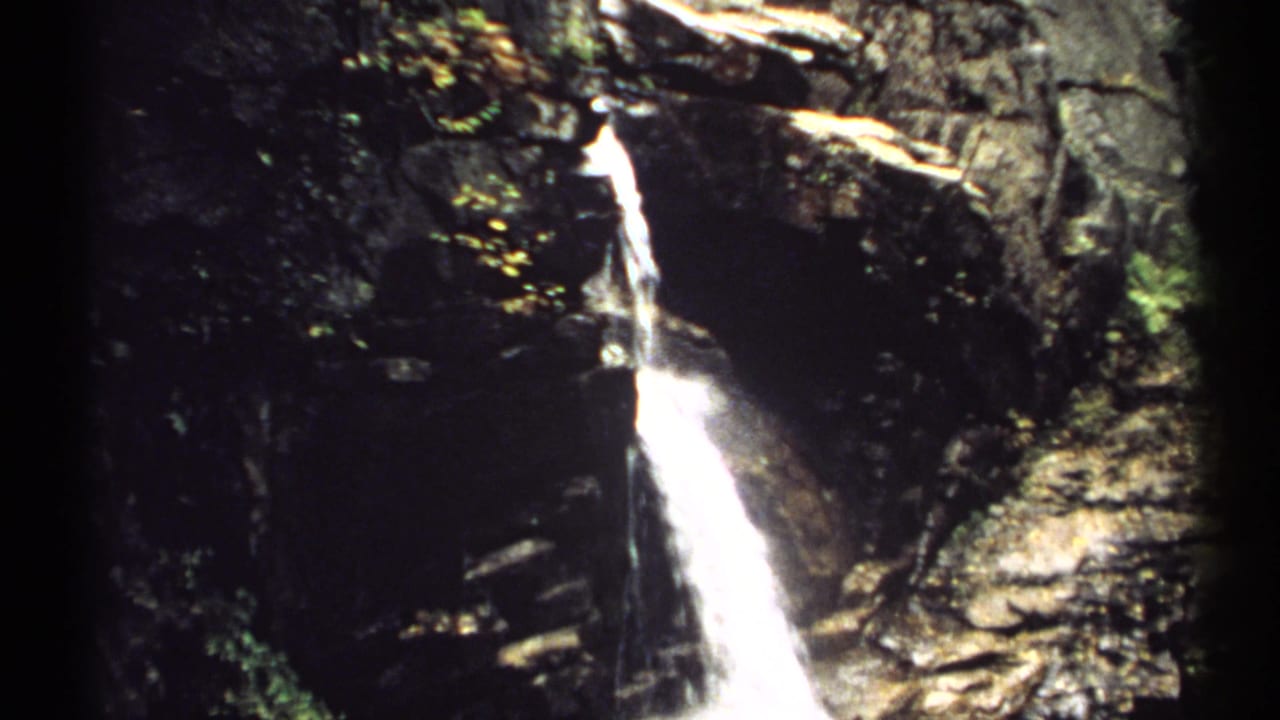 1971 New Hampshire 8mm frame showing a narrow waterfall on a dark rock face, with trees and foliage around the falls.