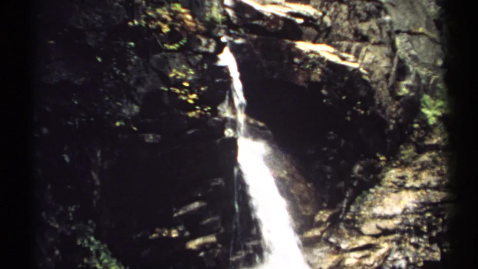 1971 New Hampshire 8mm frame showing a narrow waterfall on a dark rock face, with trees and foliage around the falls.