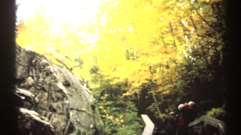1971 New Hampshire home-movie frame with yellow autumn trees, a rocky ledge, and visitors standing near boardwalk railings.