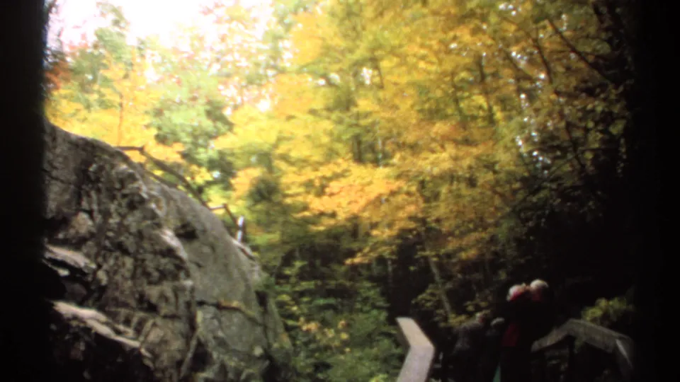 1971 New Hampshire 8mm view of a wooded rock face, autumn canopy, and distant visitors beside a boardwalk railing.