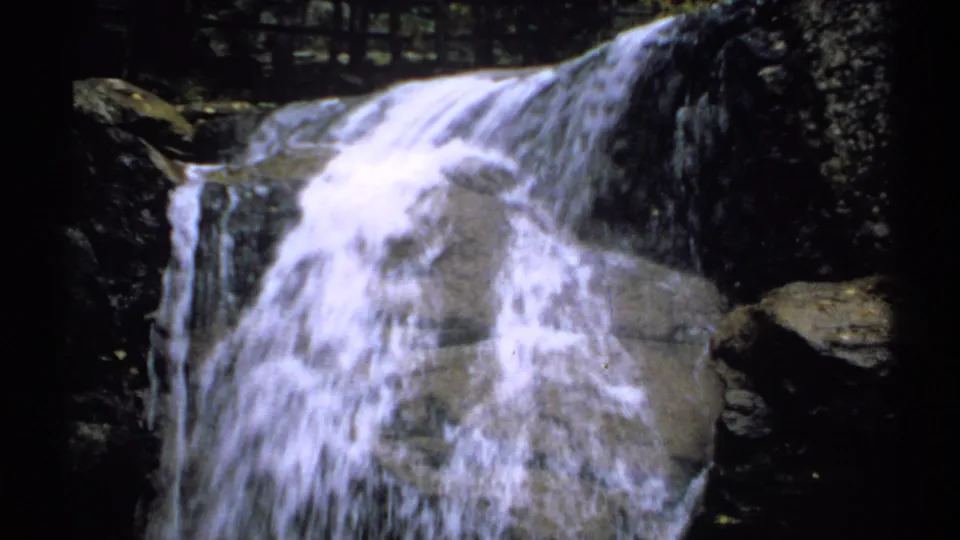 8mm home-movie frame of a small waterfall dropping over rock ledges, with dark trees and shadowed stone around it.