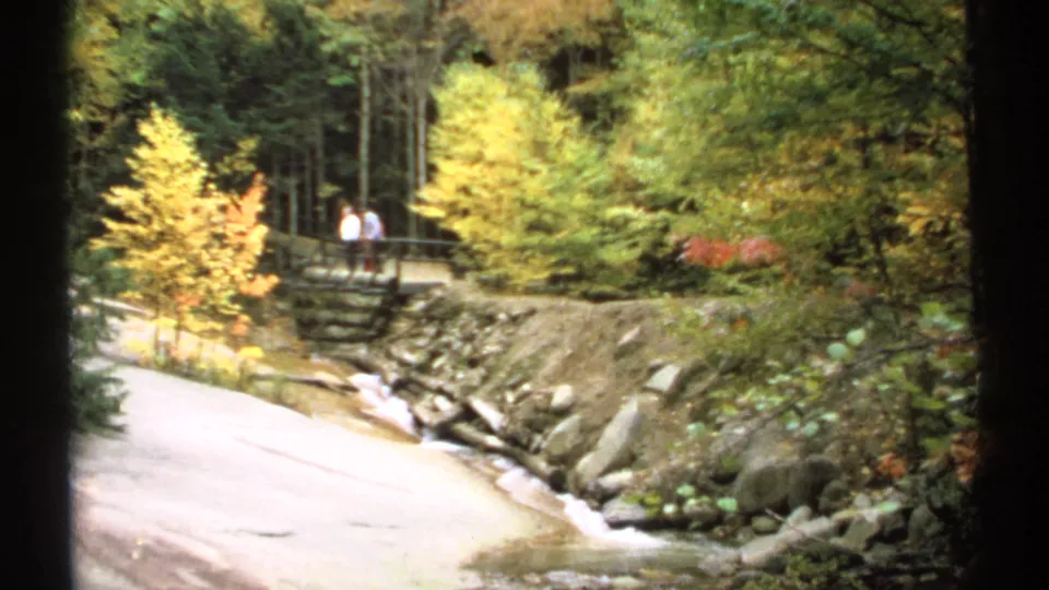 Autumn forest scene in New Hampshire with a stream, paved path, rustic bridge, and two distant people standing together.