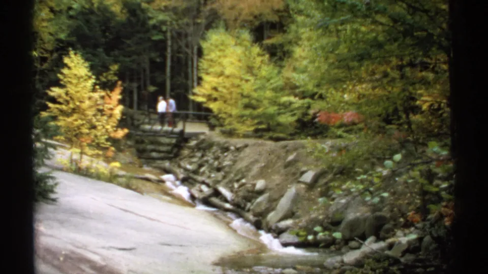 Wide home-movie view of a wooded New Hampshire stream crossing, with two distant visitors on a bridge amid autumn foliage.