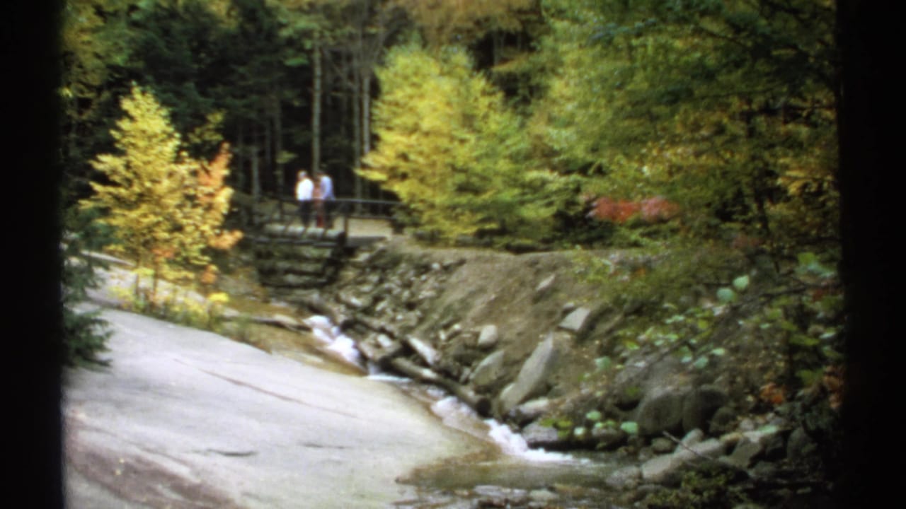 Rocky stream beside a paved path with two distant people on a wooden bridge.